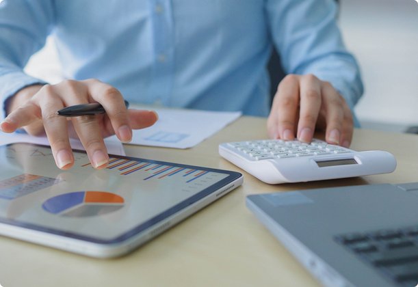 Close-up of the hands of a person in a light blue shirt working at a desk. One hand is tapping on a tablet displaying a chart, while the other hand rests on a keyboard or calculator. A laptop is partially visible in the background.