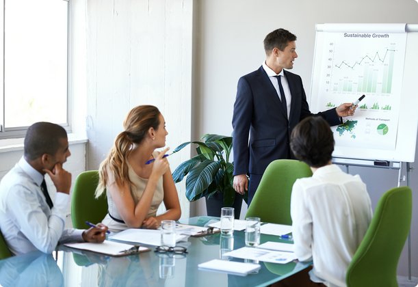A man in a suit and tie presents to a group of three listeners at a flipchart in a conference room.
