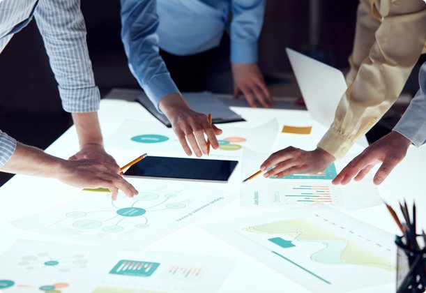 Multiple people lean over a table covered with numerous business documents featuring charts and graphs, with hands pointing to various areas, representing a collaborative analysis or discussion of data.