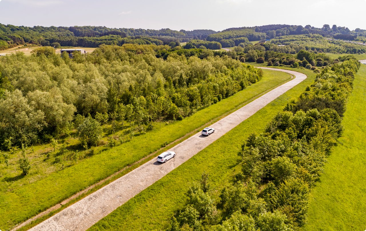 Aerial view of a long, straight asphalt road extending through a green, tree-lined landscape. Two cars are visible on the road in the distance.