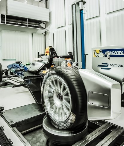 A vehicle tire with a silver rim and snow chains or measurement sensors is being tested on a rig in a brightly lit workshop hall. In the background, a blue vehicle lift and other equipment are visible.