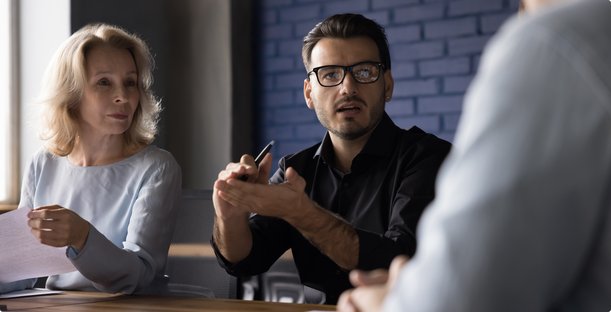 Un homme avec des lunettes et une chemise noire gesticule pendant une conversation à table, tandis qu'une femme blonde écoute attentivement et une troisième personne (vue de dos) participe également.