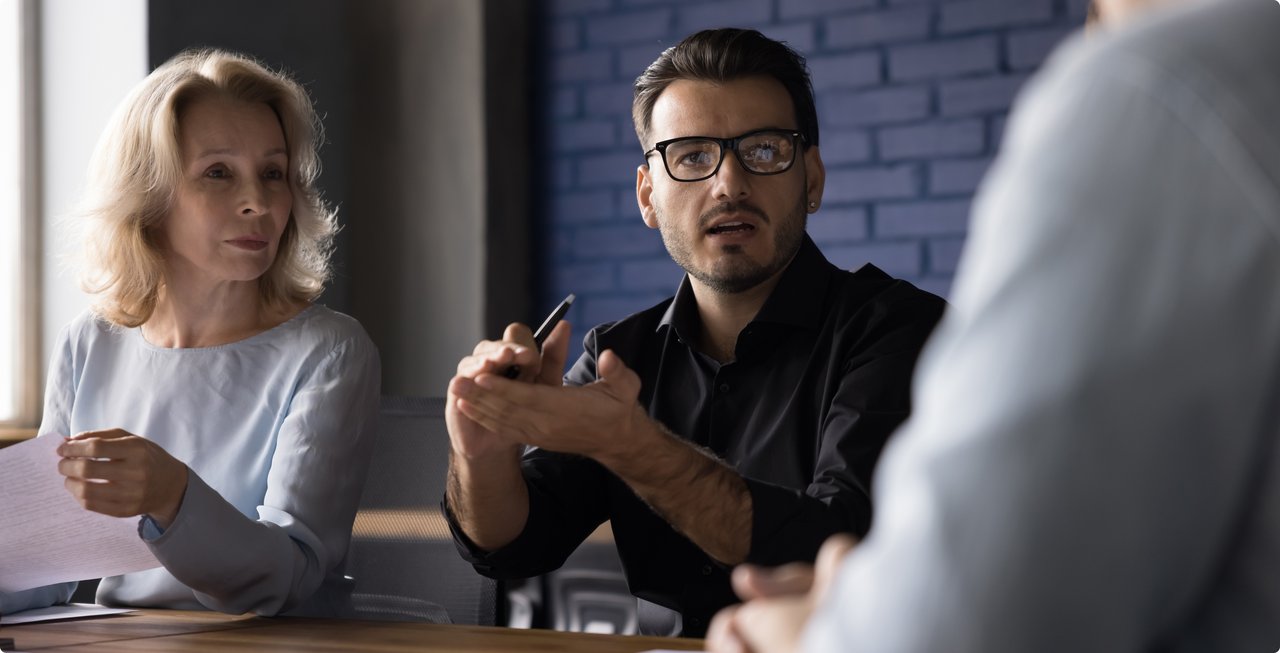A man with glasses and a black shirt gestures during a conversation at a table, while a blonde woman listens attentively and a third person (seen from behind) also participates.