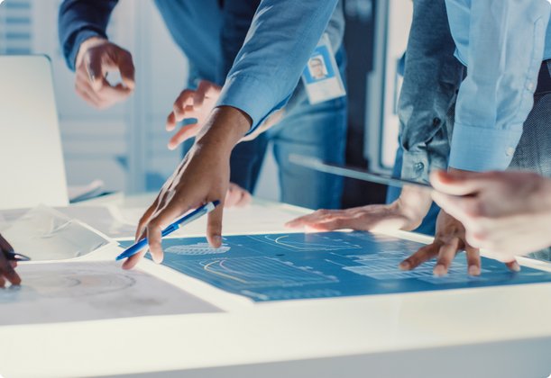 Multiple hands of people are gathered around a glowing, interactive table displaying technical blueprints or digital designs, representing a collaborative planning or design session in a modern environment.