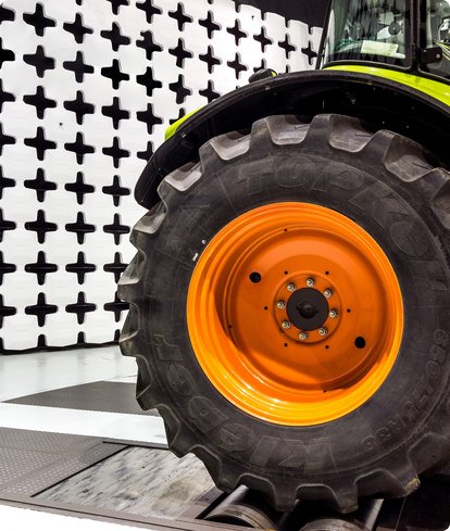 An orange wheel with off-road tires, belonging to a large vehicle, is undergoing alignment in a test bay. A black and white grid pattern is visible in the background.