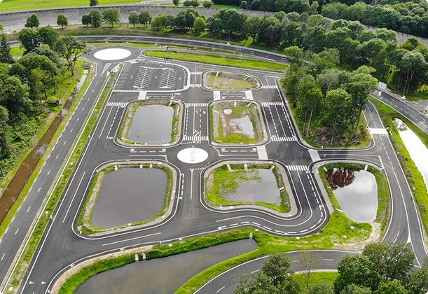 Aerial view of an extensive testing ground with various road surfaces, roundabouts, and water features, surrounded by trees and roads.