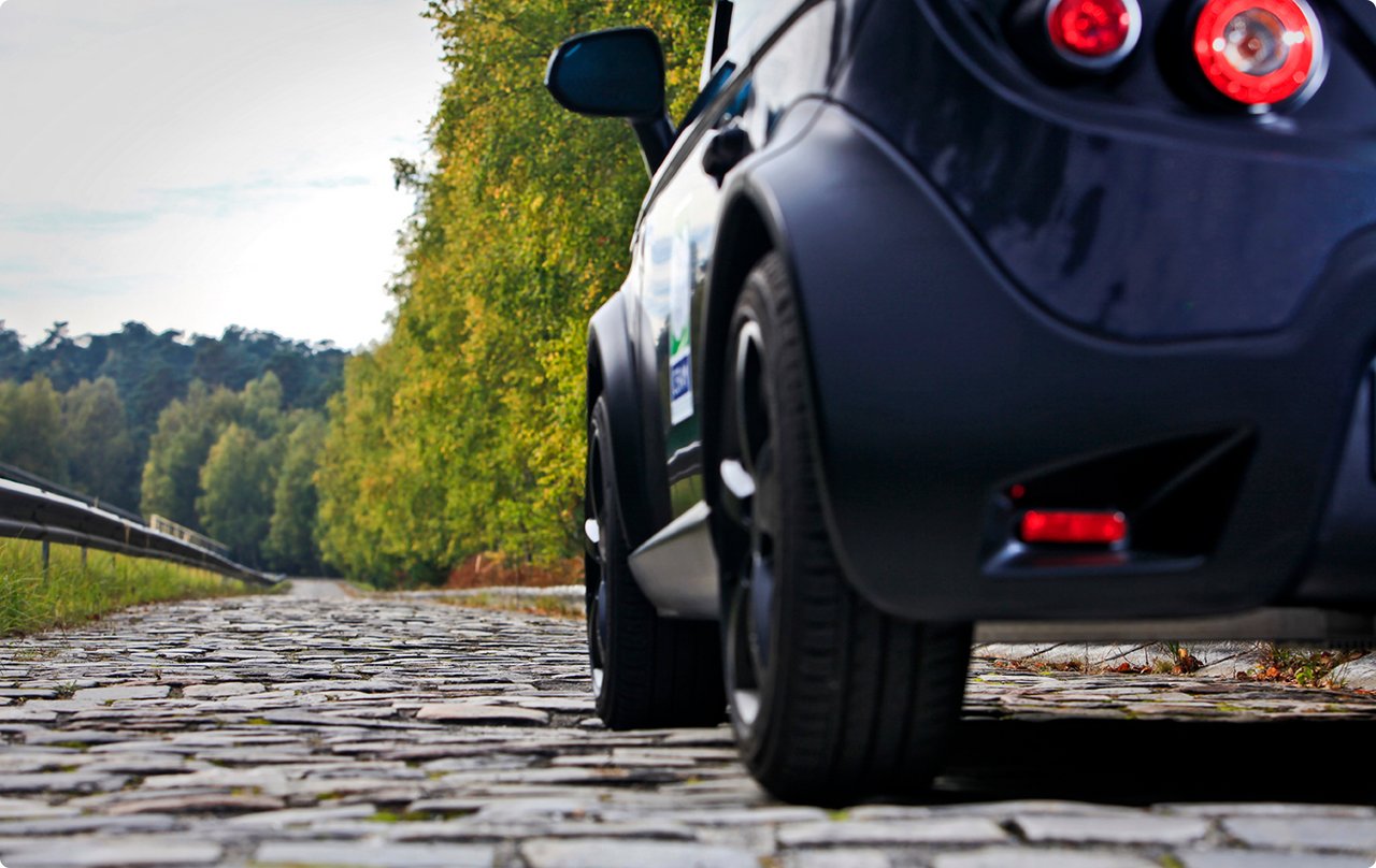 Rear view of a small, dark car parked on a rough cobblestone road. A line of trees is visible in the background.