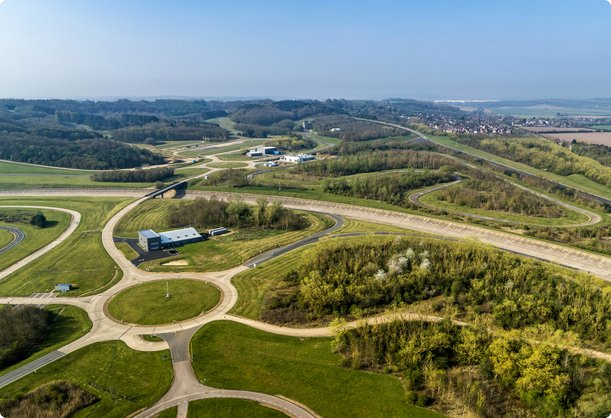 Une vaste vue aérienne présente une immense piste d'essai verte ou un terrain d'essai, composé de plusieurs routes interconnectées et de circuits ovales, entouré de forêts et de champs ouverts sous un grand ciel.