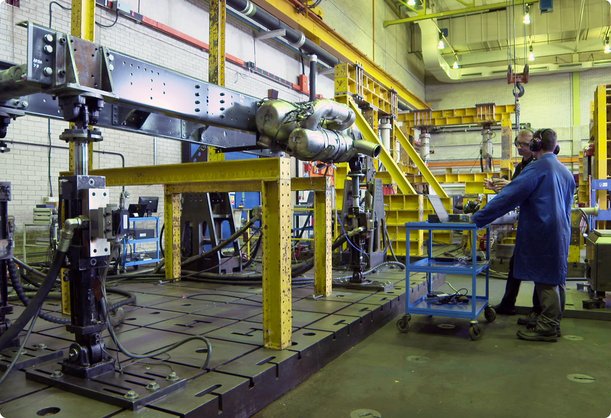 In einer großen Industrie- oder Forschungseinrichtung ist ein komplexes Maschinenteil oder ein Motor auf einem robusten Prüfstand mit gelben Stützbalken montiert. Ein Techniker in blauer Uniform steht in der Nähe und scheint Geräte auf einem Wagen zu bedienen oder zu überwachen, unter einer hohen Decke mit Deckenkränen und heller Beleuchtung.