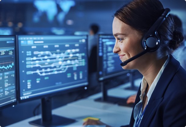 A smiling woman wearing a headset sits in front of two large computer monitors displaying complex data and diagrams. She is working in a control or command center.