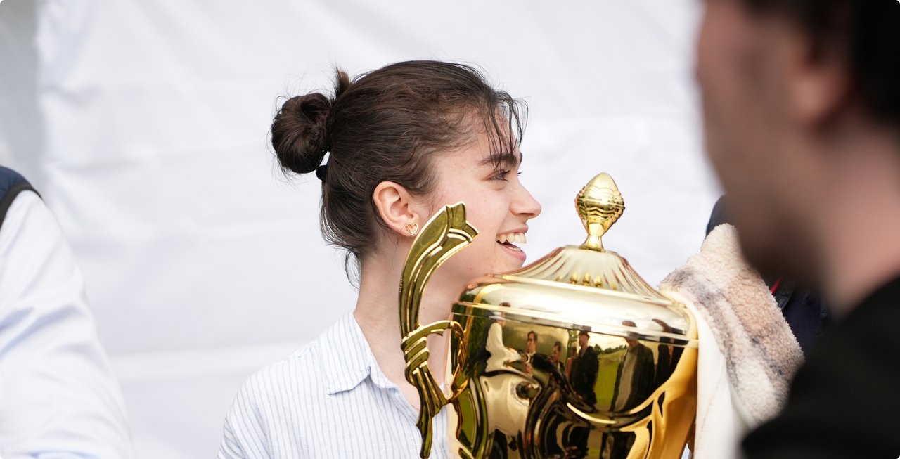 A young person with curly hair tied back is smiling brightly, holding or standing next to a large golden trophy. Part of another person's face is visible in the foreground to the right.