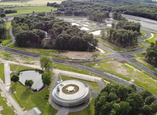 Aerial view of a large test facility with multiple roadways and water basins, surrounded by trees and green areas. In the foreground, a round building with a green roof can be seen.