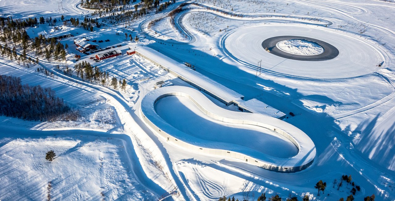A winter aerial view shows a snow-covered test track or training area with multiple oval and circular paths laid out on an icy or snowy surface.