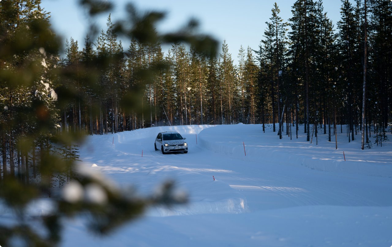 A vehicle, possibly an SUV, is driving on a snow-covered path through a forest of coniferous trees. The snow is untouched, and sunlight filters through the trees, creating a wintry and serene atmosphere.