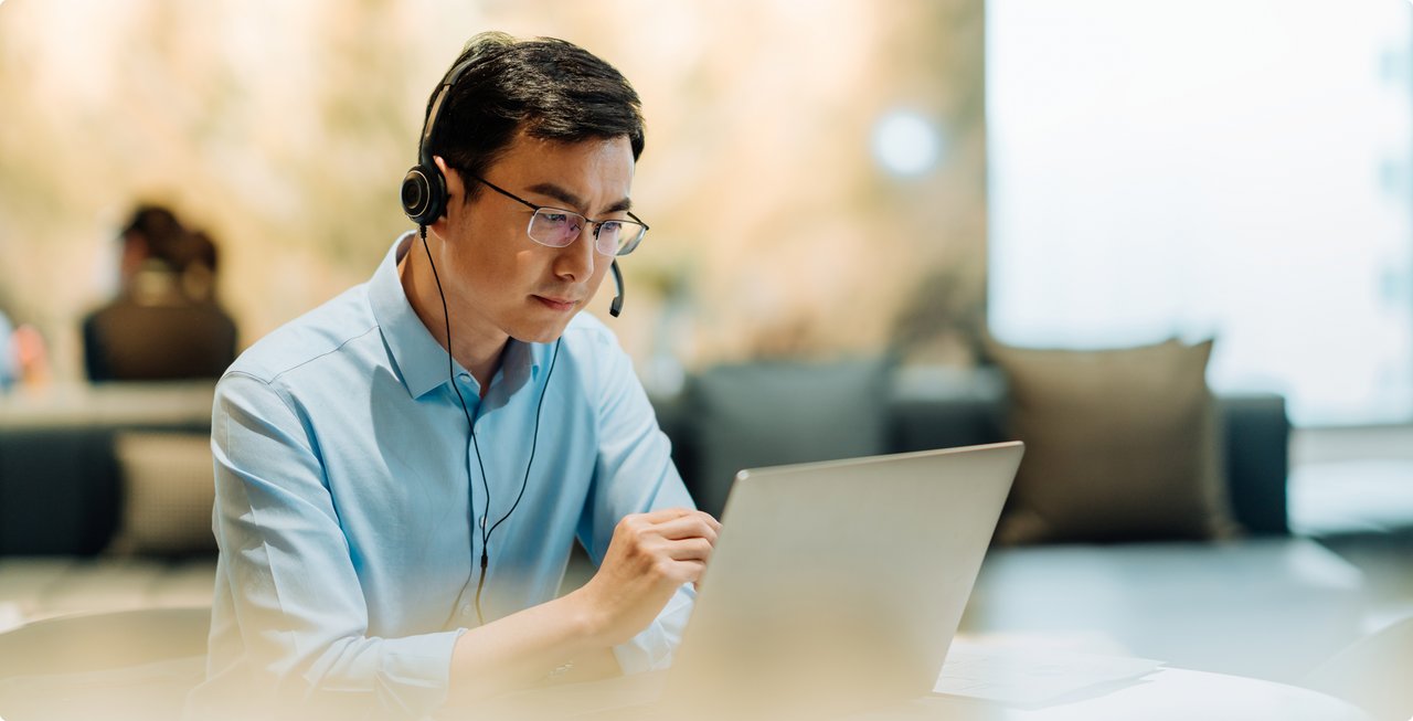 Man wearing a headset during a video call, working focused on a laptop in a relaxed environment.
