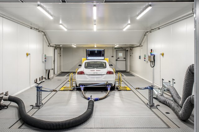 A white car is positioned on a dynamometer inside a modern, brightly lit testing chamber, with large exhaust hoses connected to its rear. The room features white walls, bright overhead lighting, and various testing equipment and sensors around the vehicle.