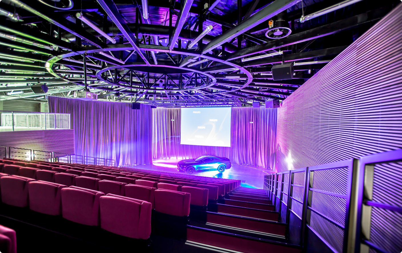 An interior view of a modern auditorium or event space with rows of red seating facing a stage. The stage features a dark, sleek car illuminated by purple and blue lights, set against a backdrop with a large screen. The walls and ceiling are adorned with intricate, swirling patterns and futuristic lighting fixtures.