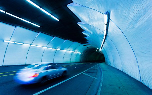 A modern car with glowing taillights drives through a futuristic-looking tunnel, illuminated with blue lights and abstract wall structures.