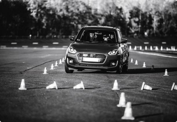 A car drives on a test track, surrounded by cones marking a course. The black and white image emphasizes the focus on the driving test.