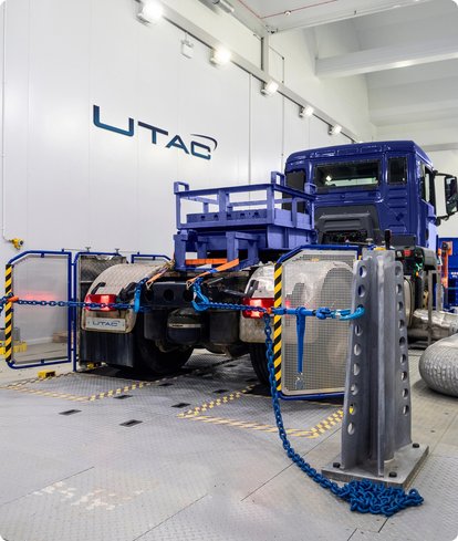 A truck cabin on a test stand inside a hall, surrounded by measurement equipment and cables, with the "WTAC" logo on the wall in the background. Debris is scattered on the floor in front of the truck.