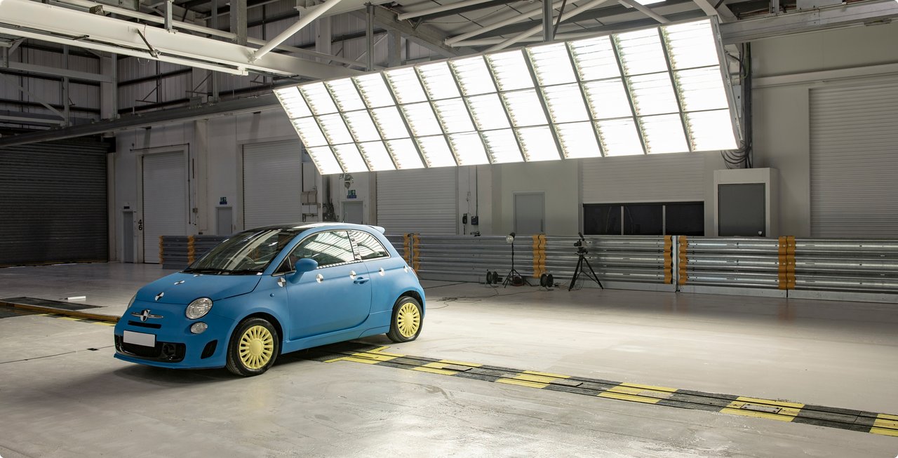A blue compact car, resembling a Fiat 500, is parked in a large testing hall. A large white lighting rig hangs above the vehicle, and barriers and other testing equipment are visible in the background.