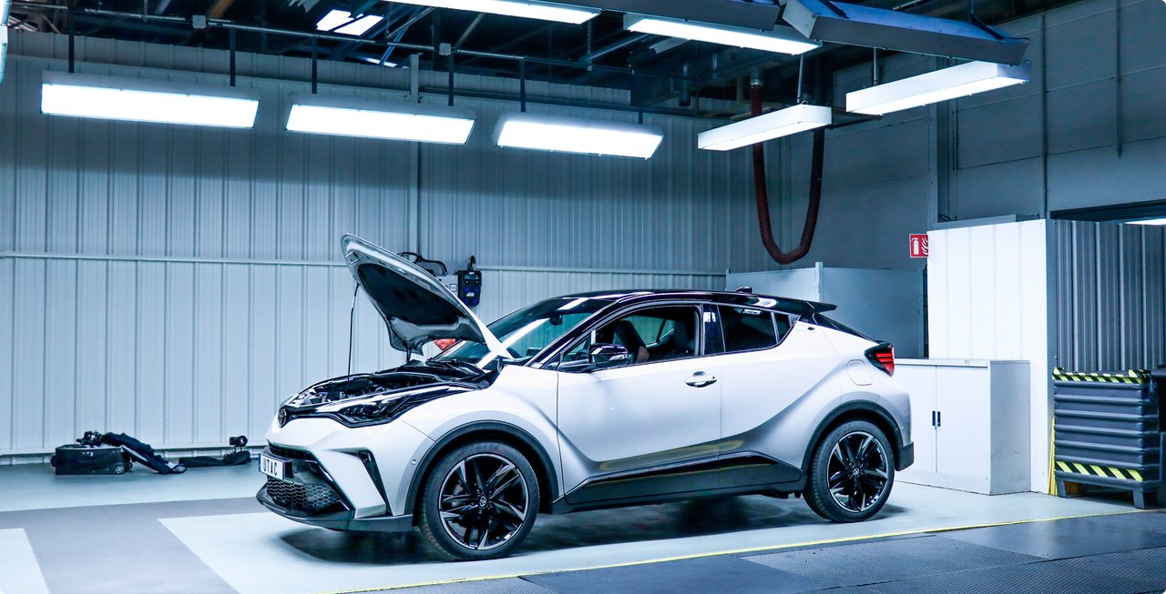 White test vehicle with open trunk in a brightly lit automotive testing environment.