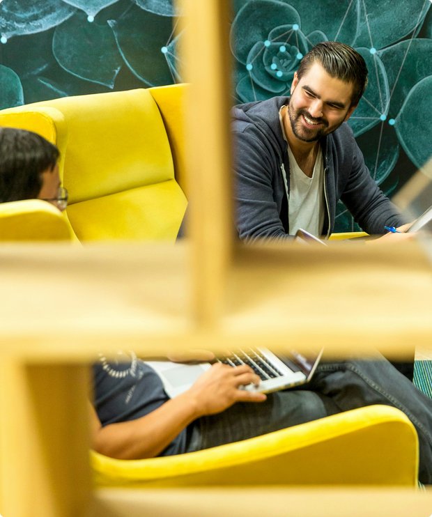 Two men chatting casually with laptops on yellow chairs – viewed through a wooden frame pattern.
