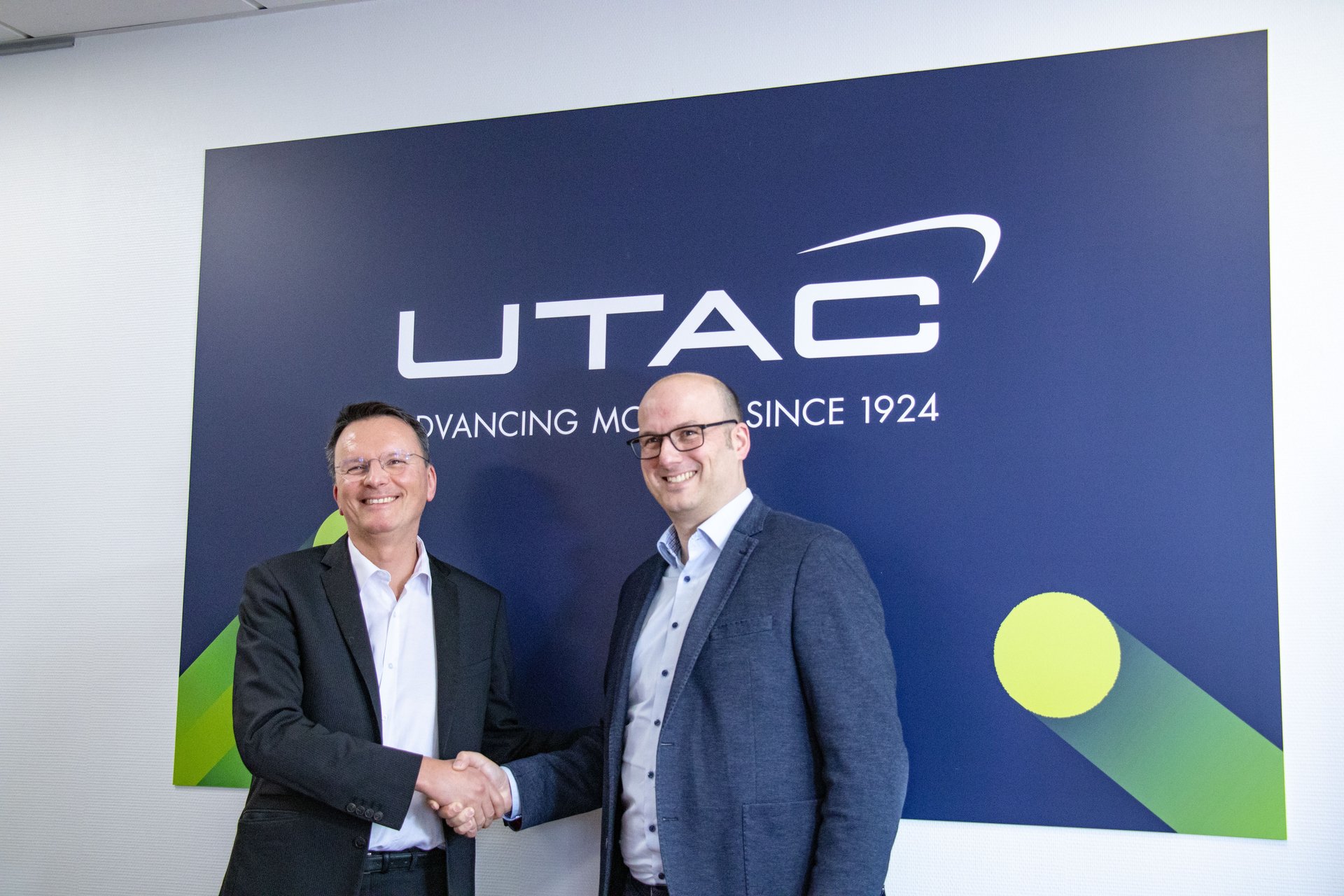 Deux hommes en tenue de ville se serrent la main devant un grand mur sombre orné du logo blanc "UTAC" et du texte en dessous. Ils sourient et regardent l'appareil photo, ce qui suggère un accord commercial ou un partenariat.