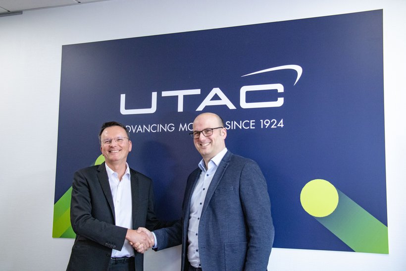 Two men in business attire are shaking hands in front of a large, dark wall with the white "UTAC" logo and text below it. They are smiling and looking at the camera, suggesting a business agreement or partnership.