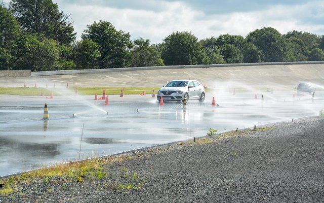 A white car drives on a wet test track marked by cones, with water splashing from its tires.