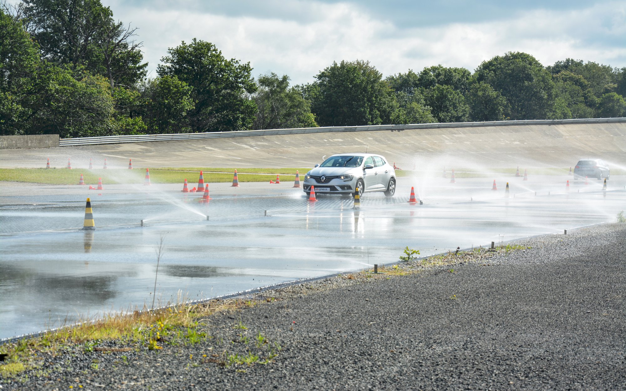 A white car drives on a wet test track marked by cones, with water splashing from its tires.