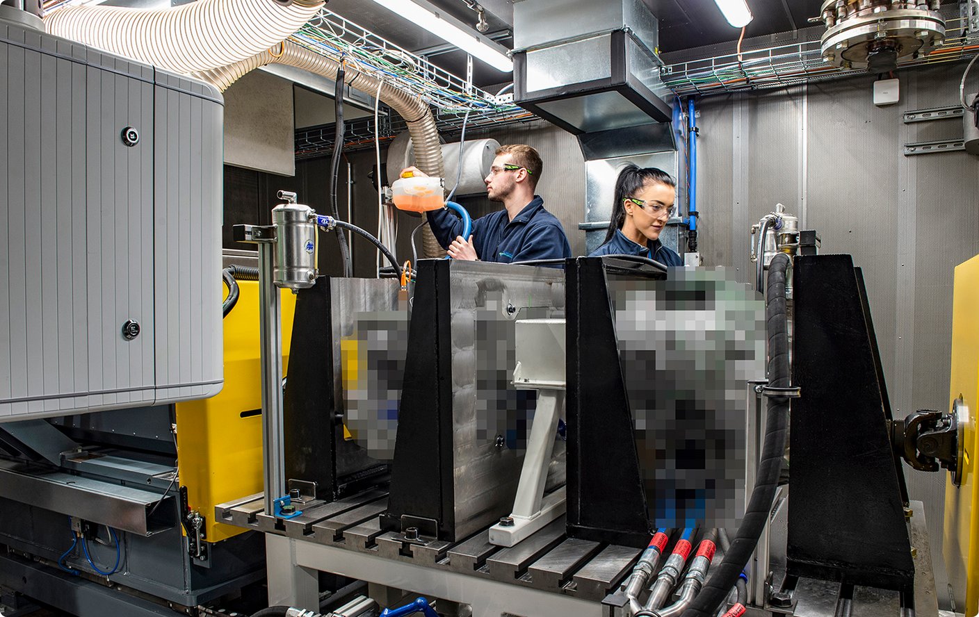 Two technicians wearing safety glasses are working in an industrial facility or laboratory, surrounded by complex machinery and cables.