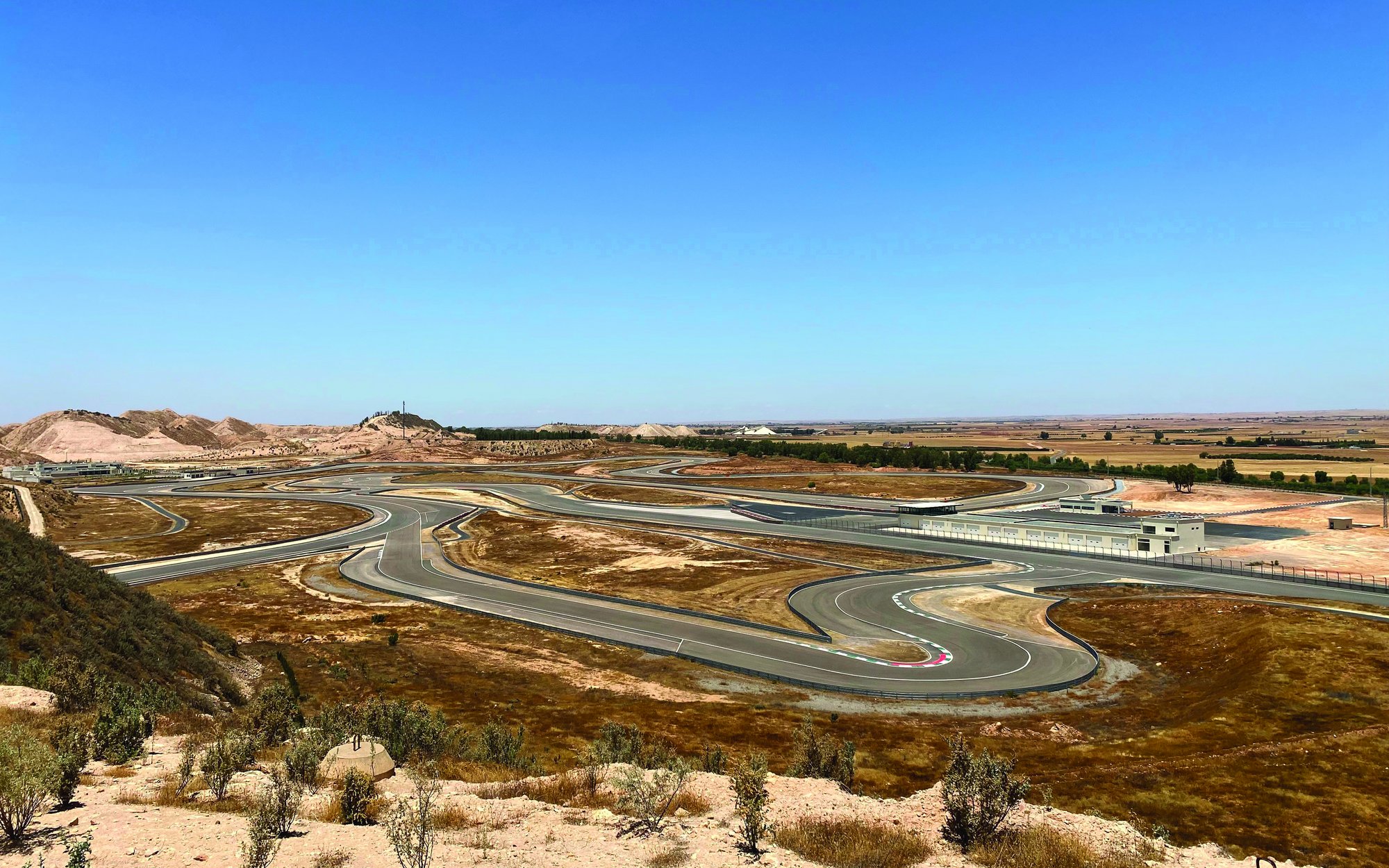 A wide landscape shot of a winding road or race track curving through a dry, rocky, and sparsely vegetated hilly terrain. In the background, more buildings and a wide, flat plain are visible under a clear blue sky.