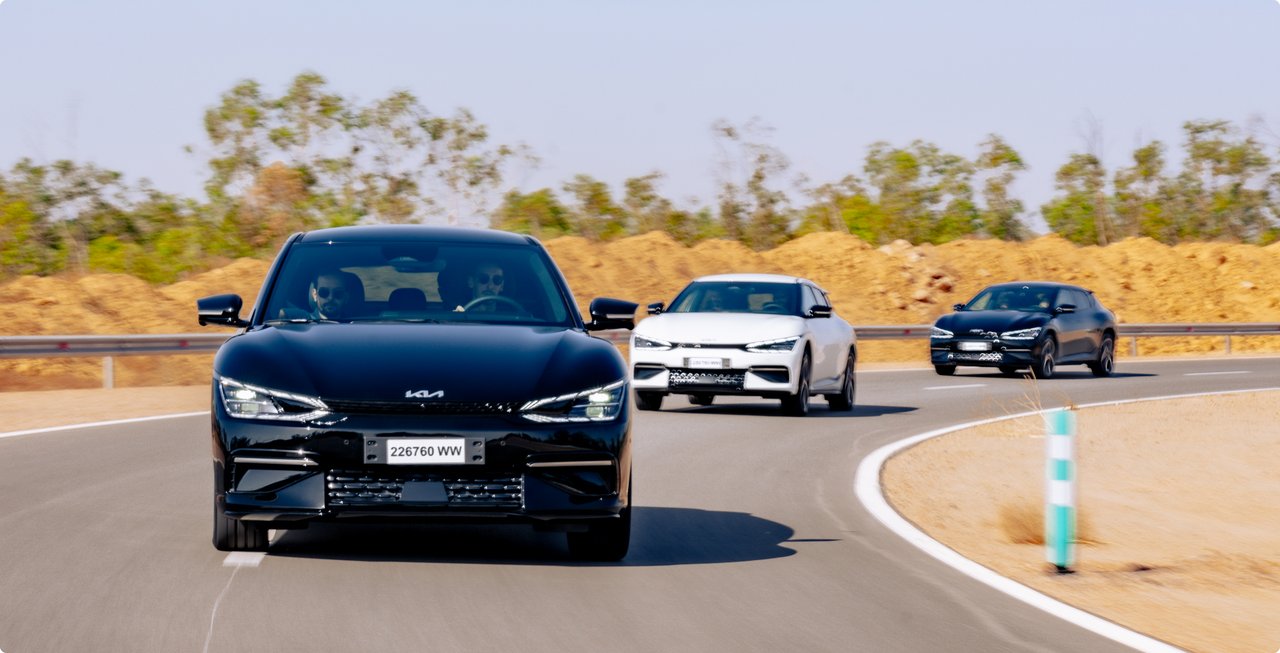 Three vehicles, led by a black SUV in the foreground, are driving on a curved test track in a sunny, dry, and barren landscape.