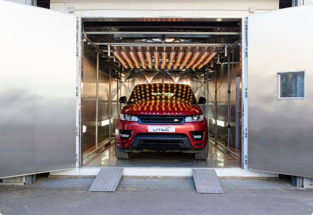 A red SUV, possibly a Range Rover, is positioned inside a large, specialized testing chamber with its doors wide open. The interior of the chamber features numerous overhead lights, possibly for simulating various environmental conditions, and the car's surface reflects these lights.