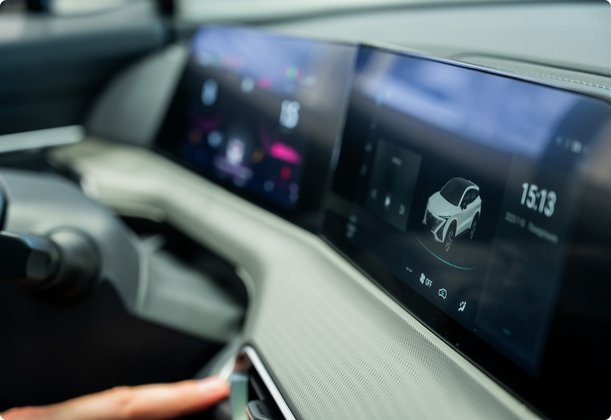 Interior view of a modern car with a large digital dashboard and infotainment system displaying a globe and other symbols. A hand is pressing a button or switch.