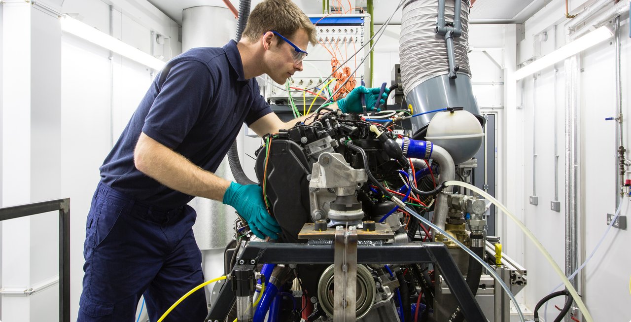 A bearded technician with glasses, wearing a dark blue work suit and turquoise gloves, working on a complex machinery setup or engine test bench in a laboratory or test room.