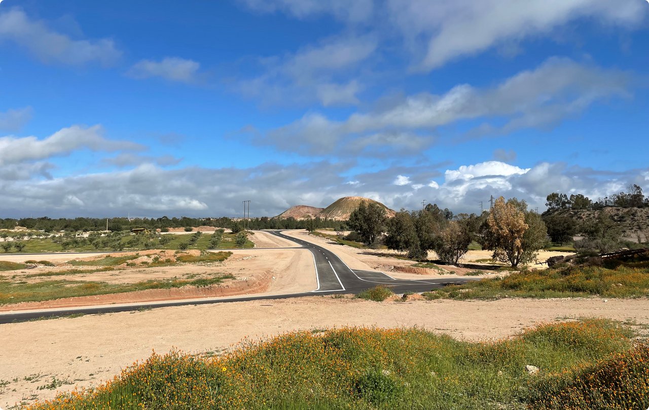 A wide, unpaved road or path leads through a dry, sandy or earthy landscape covered with low vegetation. Some buildings or structures are visible on the horizon under a cloudy, blue sky.