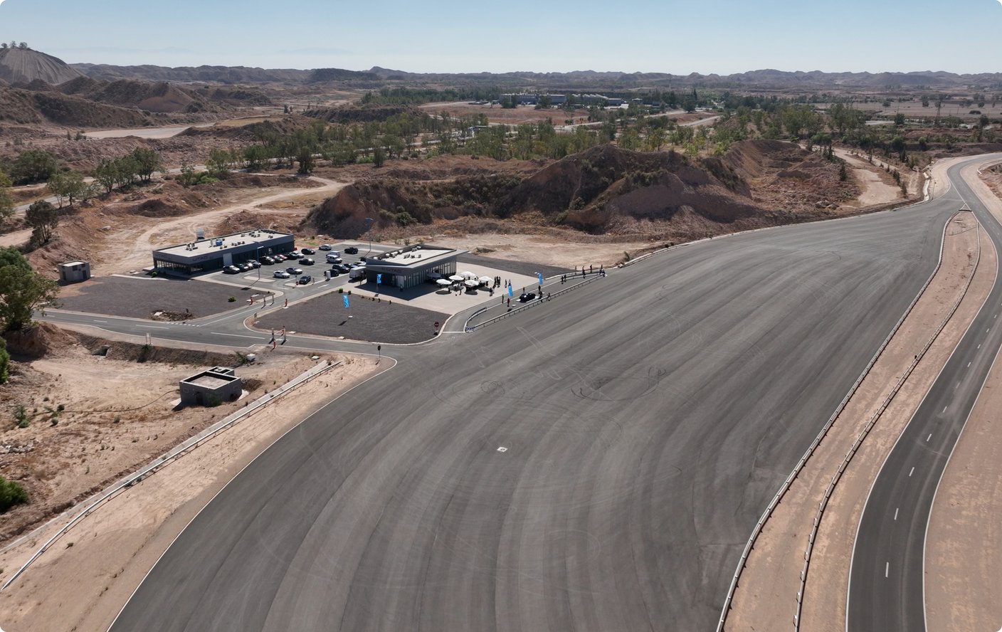 An aerial view of a newly paved road winding through a dry, arid landscape with sparse vegetation and rocky hills in the background. Two large, temporary-looking structures are visible on a gravel lot beside the road, suggesting a construction or testing site.