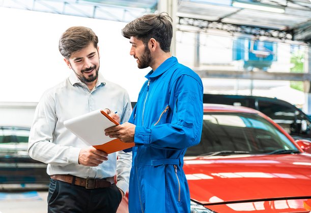 Two men in a car workshop, one in overalls and the other in a shirt, are talking and holding clipboards. A red car is visible in the background.
