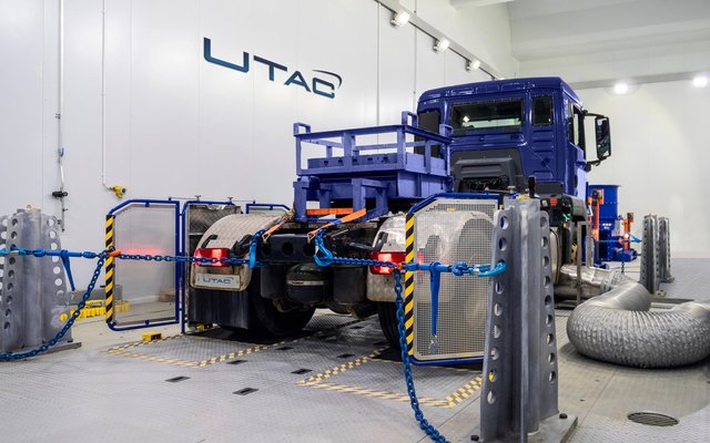 A large blue truck is being tested on a test bench in a hall, surrounded by measurement equipment, cables, and safety barriers. The "UTAC" logo is visible in the background.