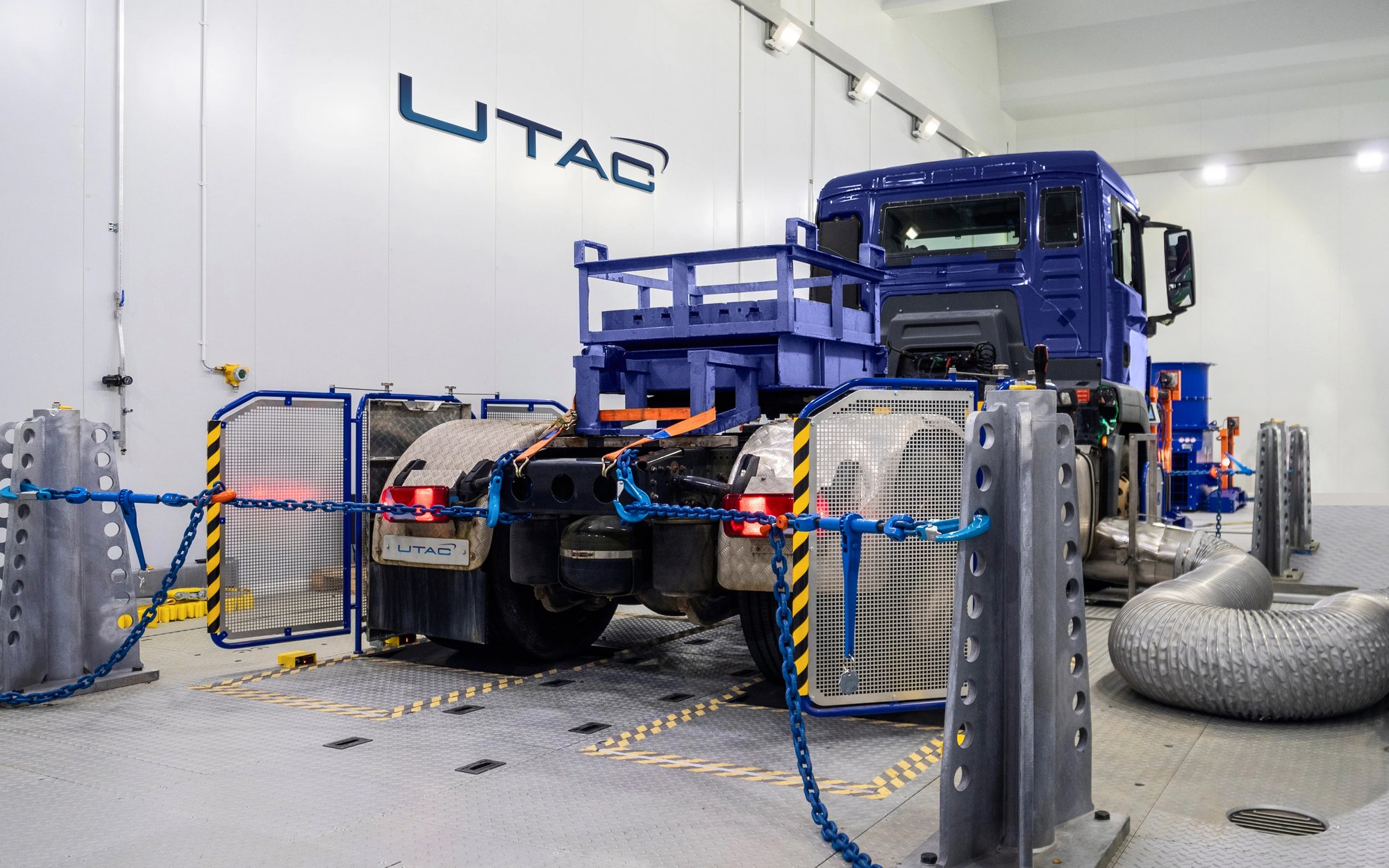 A large blue truck is being tested on a test bench in a hall, surrounded by measurement equipment, cables, and safety barriers. The "UTAC" logo is visible in the background.