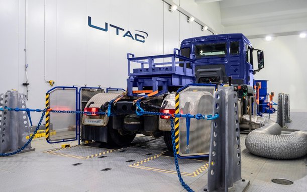 A large blue truck is being tested on a test bench in a hall, surrounded by measurement equipment, cables, and safety barriers. The "UTAC" logo is visible in the background.