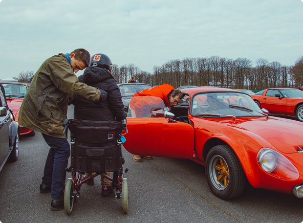 A man leans towards a person in a wheelchair, who is wearing warm clothing, while a striking red vintage sports car with an open door is parked nearby. It appears to be an outdoor event on a cool day.