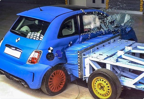 A blue compact car, resembling a Fiat 500, is impacted by a mobile sled with a deformable block in a crash test. The impact occurs at the rear of the vehicle, causing the rear window to shatter and debris to fly.