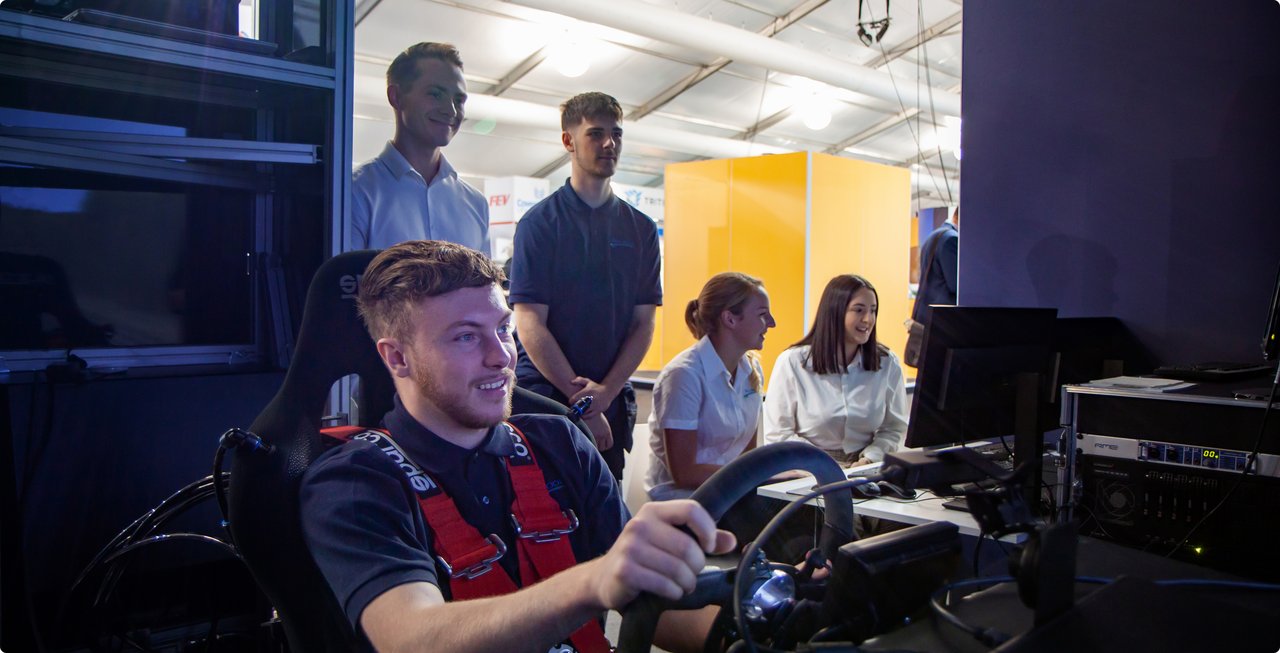 A young man wearing a red harness sits in a driving simulator, holding a steering wheel, while three other people watch in the background.