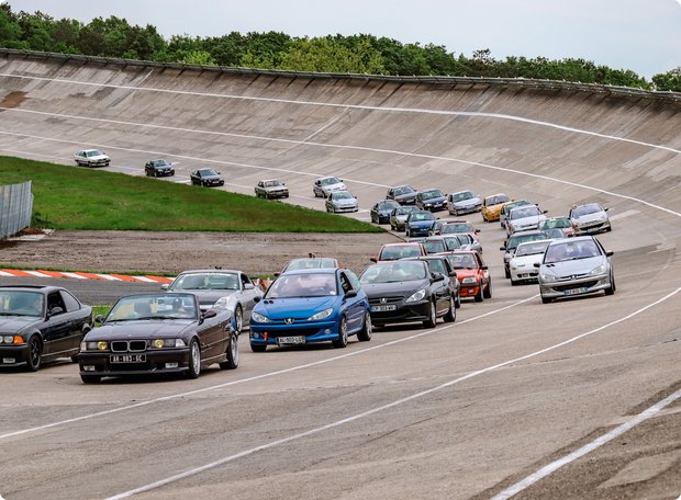 A line of classic and modern cars, including a prominent blue car, drives along a historic, banked oval race track or test track. The track features a worn concrete surface with painted dashed lines, and a grassy infield with more cars visible in the distance.