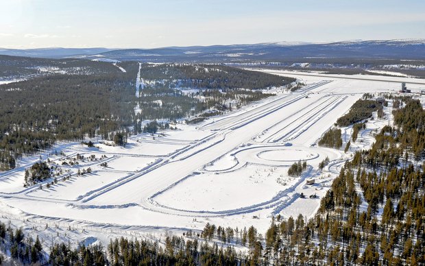 An aerial view of a vast snow-covered landscape, possibly a winter test track or a ski resort, with multiple parallel tracks cut into the snow, winding trails, and dense evergreen forests bordering the open areas.