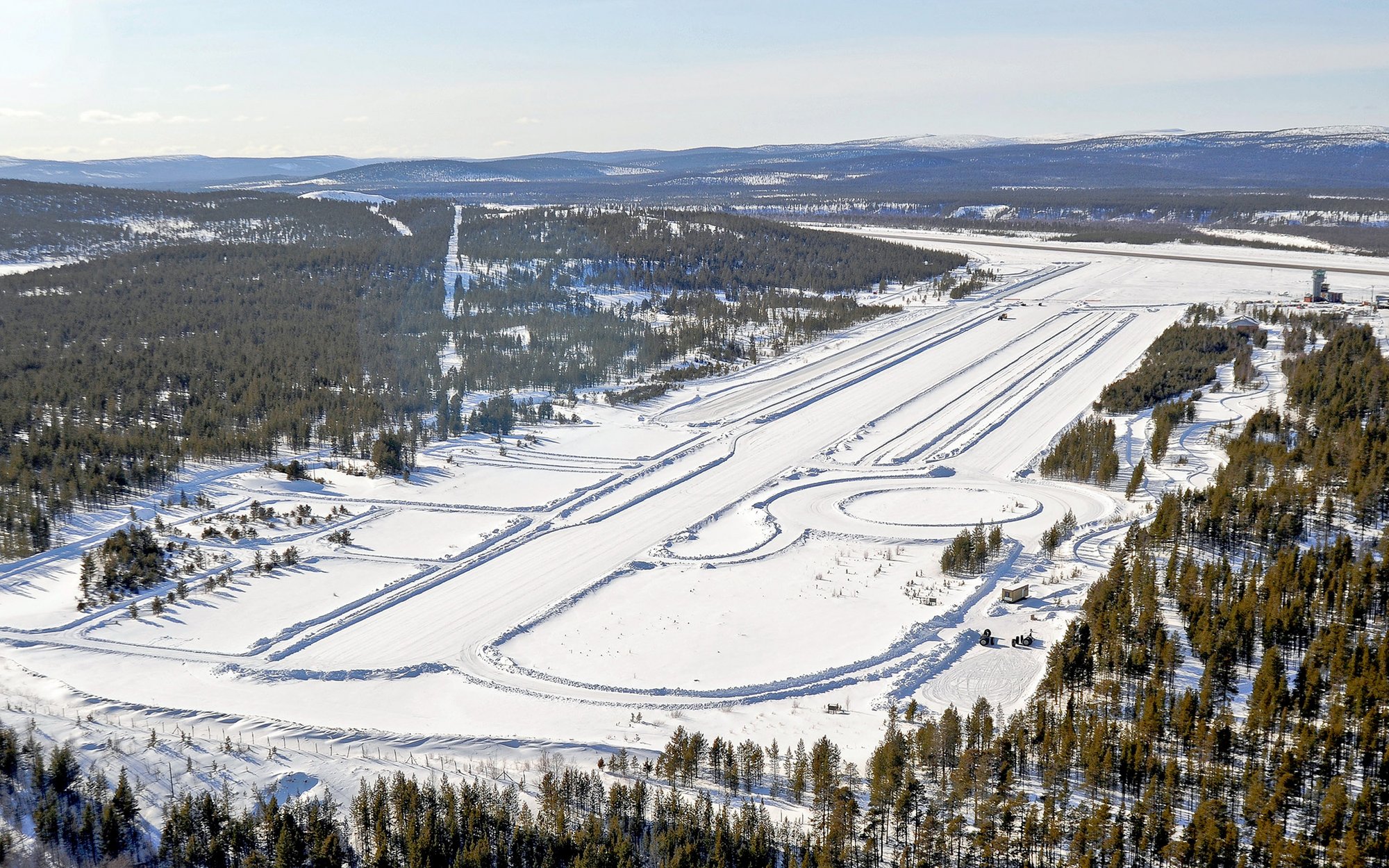 An aerial view of a vast snow-covered landscape, possibly a winter test track or a ski resort, with multiple parallel tracks cut into the snow, winding trails, and dense evergreen forests bordering the open areas.