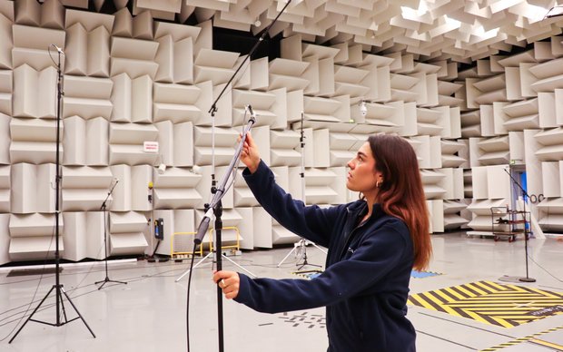 A female technician with long brown hair adjusting a microphone on a stand in an anechoic chamber. The walls and ceiling of the room are covered with grey, sound-absorbing wedges.